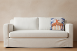 A square decorative throw pillow featuring an illustration of two giraffes, styled on an olive green velvet sofa in a modern living room setting.