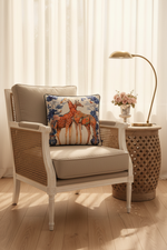 A square decorative throw pillow featuring an illustration of two giraffes on a white background with blue floral details, styled on an olive green velvet sofa next to a black side table.