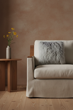 A beige upholstered armchair with a textured gray faux-fur throw pillow, set in a minimalist room with a warm brown plaster wall, light wood floor, and a small wooden side table holding yellow flowers.