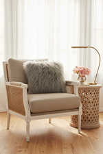 A beige upholstered armchair with woven cane sides and a fluffy gray throw pillow, styled in a bright room next to a wooden side table with a lamp and flowers.