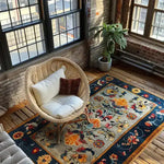 Colorful Carnival Hand Tufted Wool Rug in a cozy living room setting. The large area rug features vibrant floral patterns in rich colors, adding warmth to the wooden floor. A comfortable wicker chair with cushions sits atop the rug, enhancing the inviting atmosphere. Ideal for those seeking stylish area rugs for the living room.