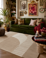 A styled living room featuring a large abstract area rug in beige and cream tones, a deep green velvet sofa with patterned pillows, and a decorative side table in the foreground.