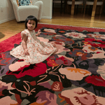 Child in a floral dress sitting on a colorful floral-patterned rug in a room with wooden floors and furniture.