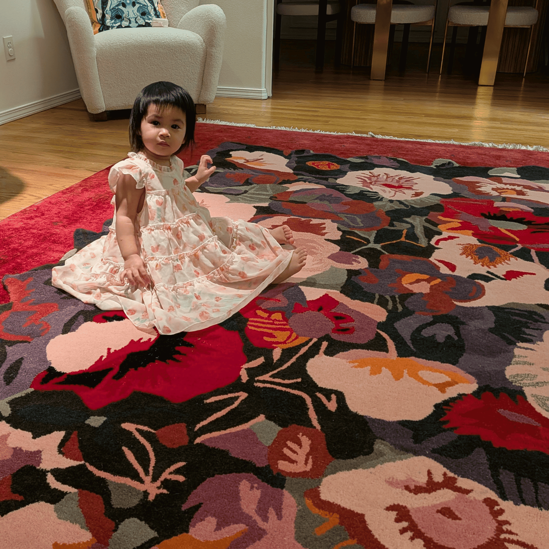 Child in a floral dress sitting on a colorful floral-patterned rug in a room with wooden floors and furniture.