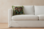 A cream-colored fabric sofa with a single decorative throw pillow featuring a colorful embroidered tiger and tropical foliage pattern with green fringe, set against a textured beige wall and light wood flooring.