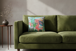 A square decorative throw pillow with a colorful tropical and zebra print sits on an olive green velvet sofa. The pillow features a turquoise background with pink and white zebras, palm trees, and a beach hut. The sofa is styled in a modern living room setting with a black side table and vase on the left.