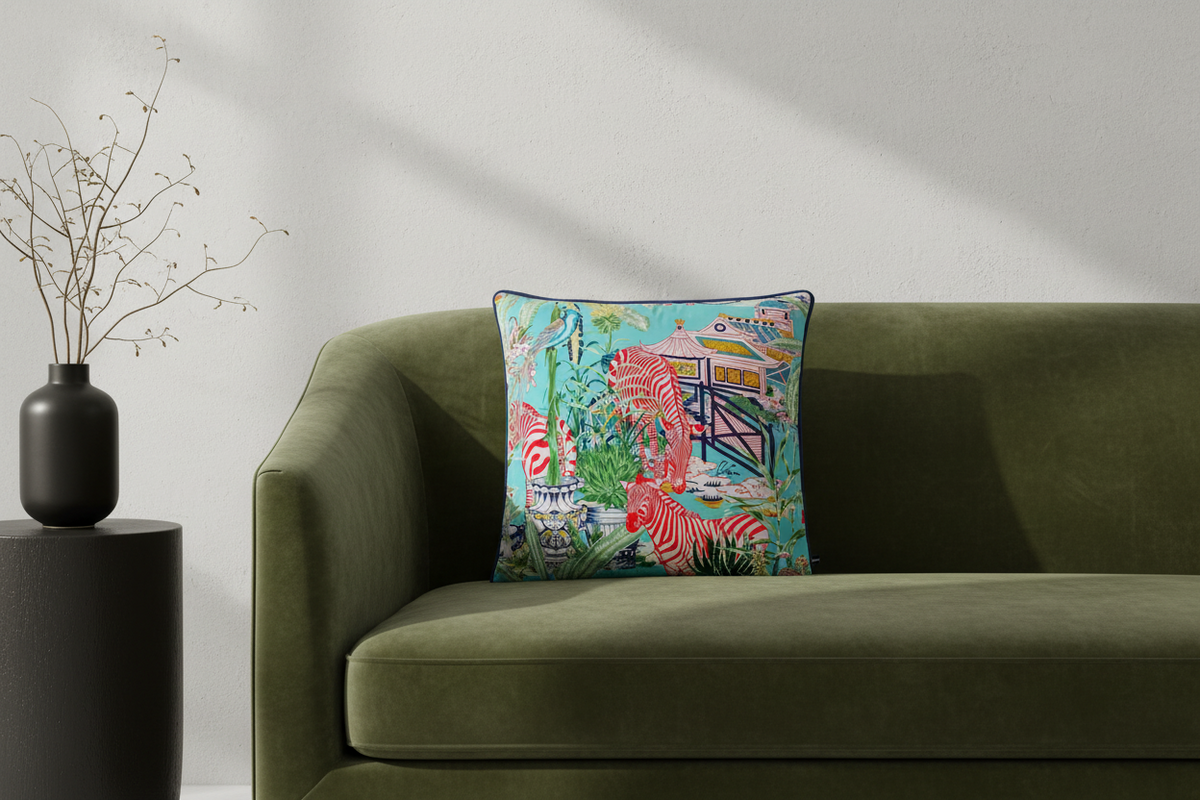 A square decorative throw pillow with a colorful tropical and zebra print sits on an olive green velvet sofa. The pillow features a turquoise background with pink and white zebras, palm trees, and a beach hut. The sofa is styled in a modern living room setting with a black side table and vase on the left.