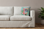 A square decorative throw pillow with a colorful tropical and zebra print sits on an olive green velvet sofa. The pillow features a turquoise background with pink and white zebras, palm trees, and a beach hut. The sofa is styled in a modern living room setting with a black side table and vase on the left.