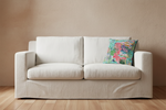 A light beige upholstered loveseat with two seat cushions and a colorful tropical floral throw pillow, set against a textured beige wall and light wood floor.