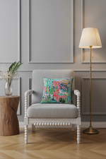 A square decorative throw pillow with a colorful tropical and zebra print sits on an olive green velvet sofa. The pillow features a turquoise background with pink and white zebras, palm trees, and a beach hut. The sofa is styled in a minimalist living room setting with a black vase of dried branches on a side table.