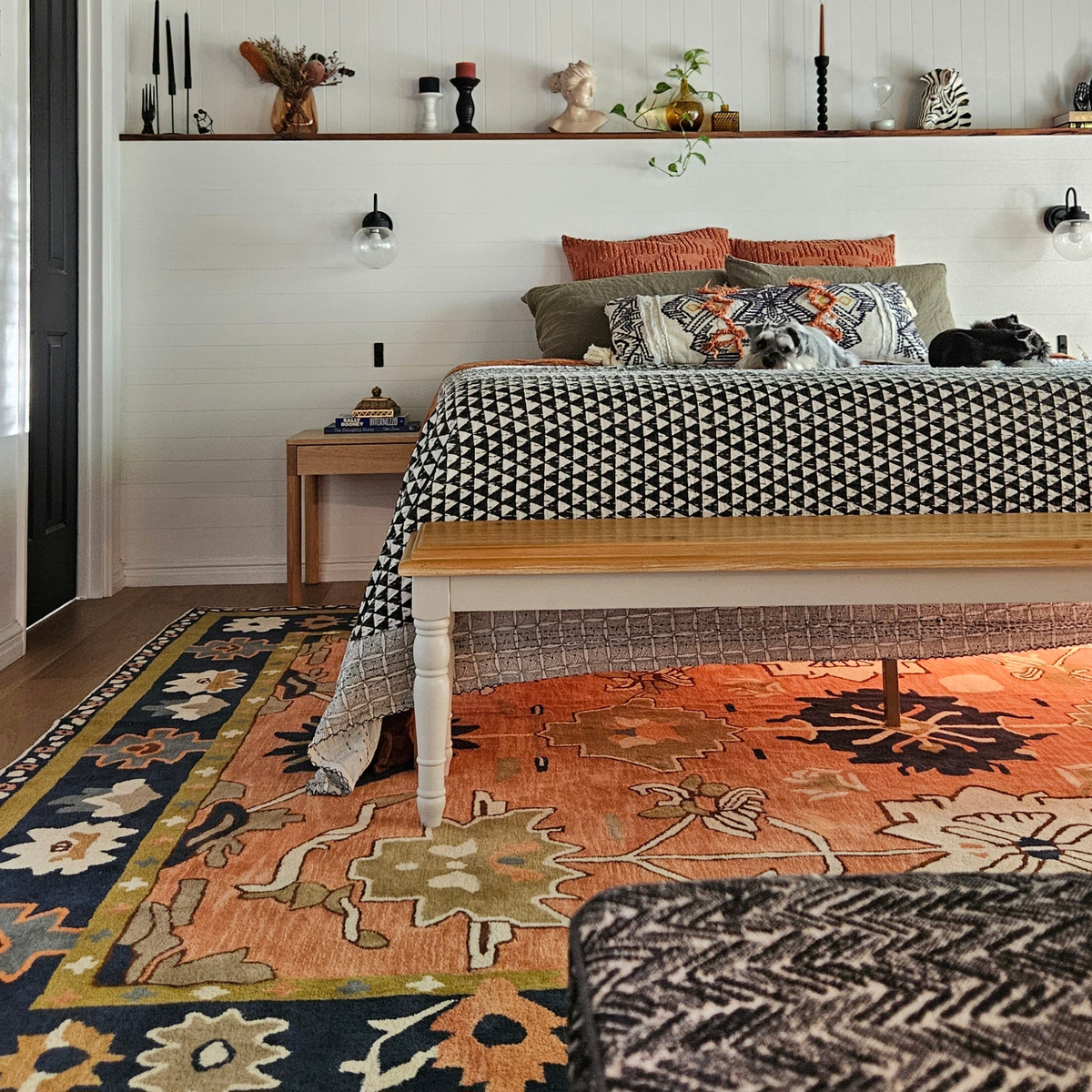 A styled bedroom scene featuring a large, ornate area rug with a warm orange, beige, and navy floral pattern. The rug is styled under a wooden bed frame with patterned bedding, against a white shiplap wall with decorative wall sconces and a shelf.