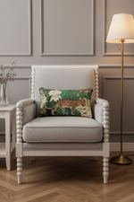 A light gray upholstered armchair with turned wooden arms and legs, styled in a room with a green leopard-print throw pillow, a floor lamp, and a side table.