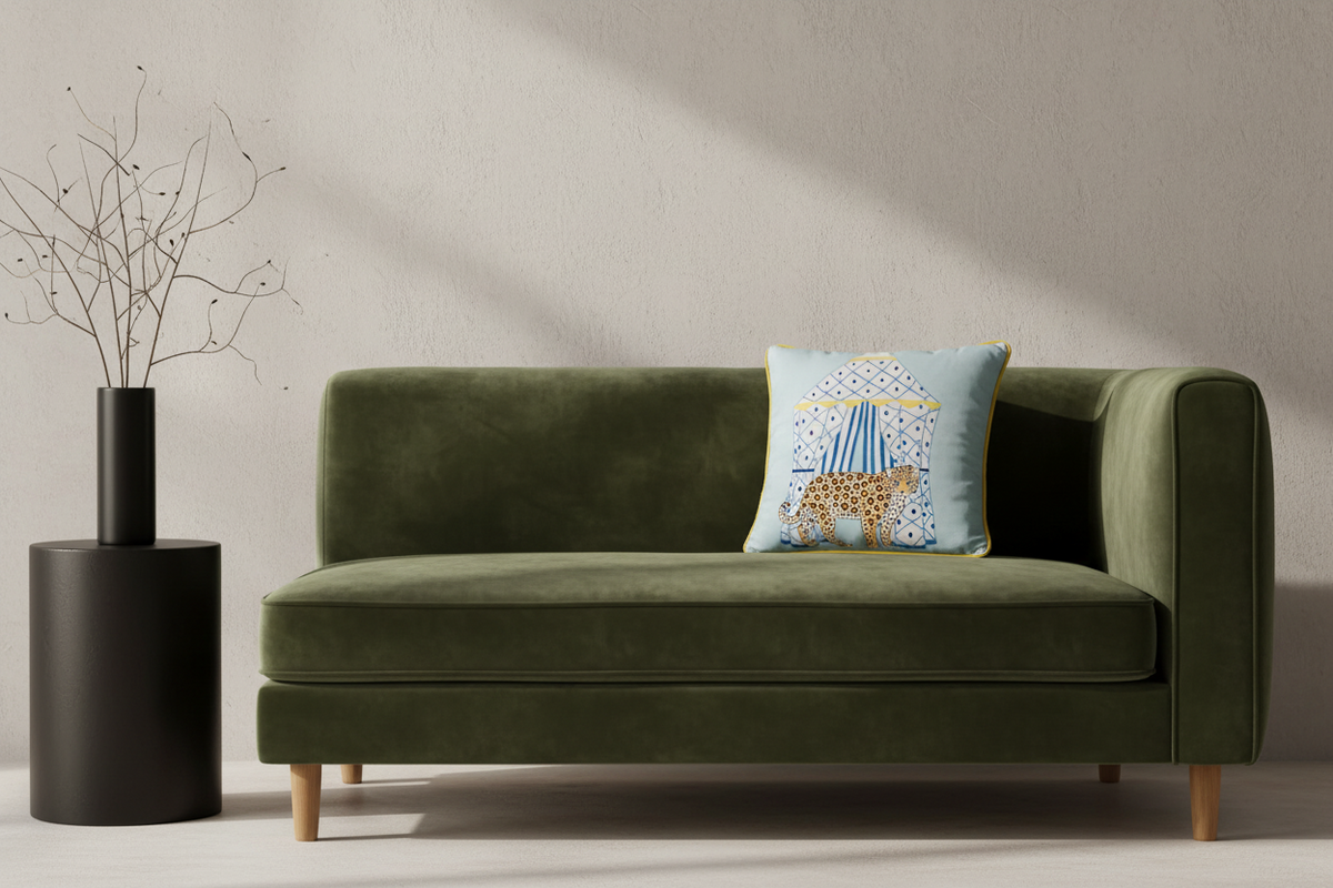 An olive green velvet loveseat sofa with light wood legs, styled with a light blue throw pillow featuring a giraffe under a circus tent, next to a black cylindrical side table with dried branches.