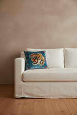 A square decorative throw pillow featuring a roaring tiger design on a teal background with red floral accents, styled on a cream-colored slipcovered sofa against a textured beige wall and light wood flooring.