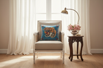 A decorative square throw pillow featuring a vibrant blue background with a detailed tiger print and red floral accents, styled on a beige armchair in a bright, sunlit room with white curtains and a wooden side table.