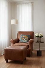 A brown leather armchair with a matching ottoman in a bright room, styled with a blue tiger-print throw pillow, a floor lamp, and a side table with white flowers.