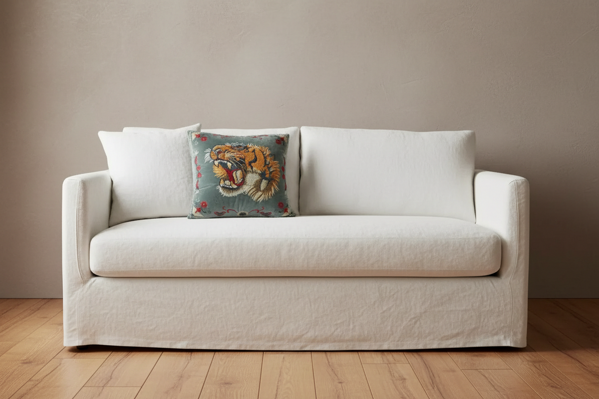 A cream-colored fabric slipcovered loveseat sofa with a single decorative square throw pillow featuring a roaring tiger design, set against a neutral beige wall and light wood floor.