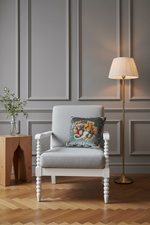 A white armchair with a light gray upholstered seat, styled in a room with gray paneled walls and a wood floor. A decorative throw pillow featuring a roaring tiger is placed on the chair, alongside a wooden side table with a vase of greenery and a brass floor lamp.