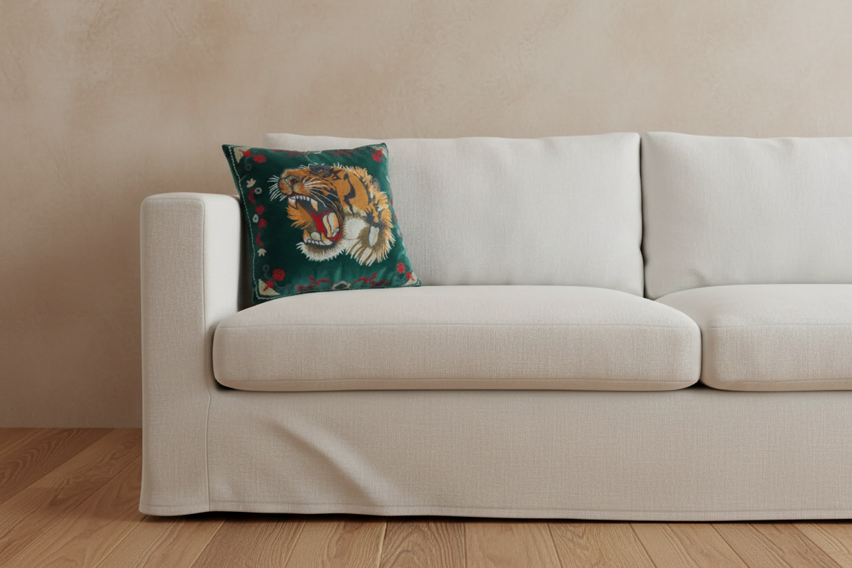 A cream-colored fabric sofa slipcover with a matching skirt, styled in a living room setting against a textured beige wall and light wood flooring, featuring a decorative green tiger throw pillow.