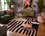 A large area rug with a bold abstract pattern in black, pink, cream, and mustard yellow, styled in a modern, colorful living room with a green sofa, pink armchair, and wooden floors.