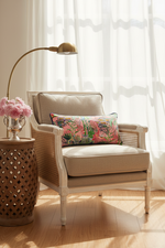 A brown leather wingback armchair with a matching ottoman in a warm, styled room. A colorful floral accent pillow rests on the chair, alongside a floor lamp and a side table with a vase of white flowers.