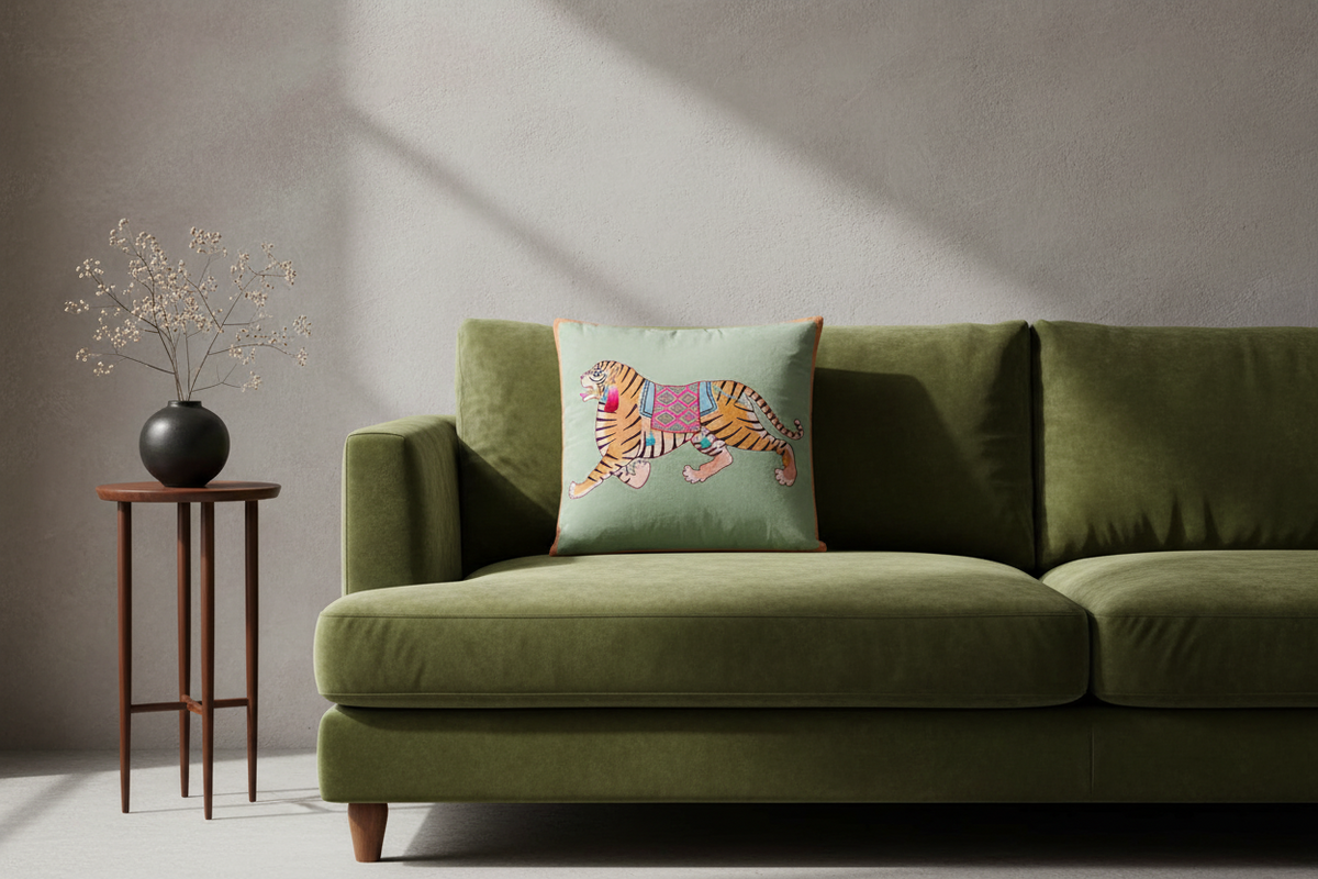 An olive green velvet sofa in a modern living room setting, featuring a square mint green throw pillow with an embroidered tiger design. A small wooden side table with a black vase of dried branches stands to the left against a textured gray wall with soft diagonal shadows.