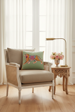 A styled living room scene featuring a beige armchair with a woven cane back, a matching beige cushion, and a decorative throw pillow with an embroidered tiger. A small wooden side table with a brass lamp and pink flowers sits next to the chair in front of sheer white curtains.