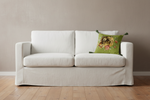 A white slipcovered two-seat sofa with a decorative green floral throw pillow, set against a neutral beige wall and light wood flooring.