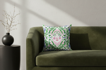 A square decorative throw pillow with a colorful folk-art face pattern, styled on an olive green velvet sofa next to a black side table with a vase of branches.