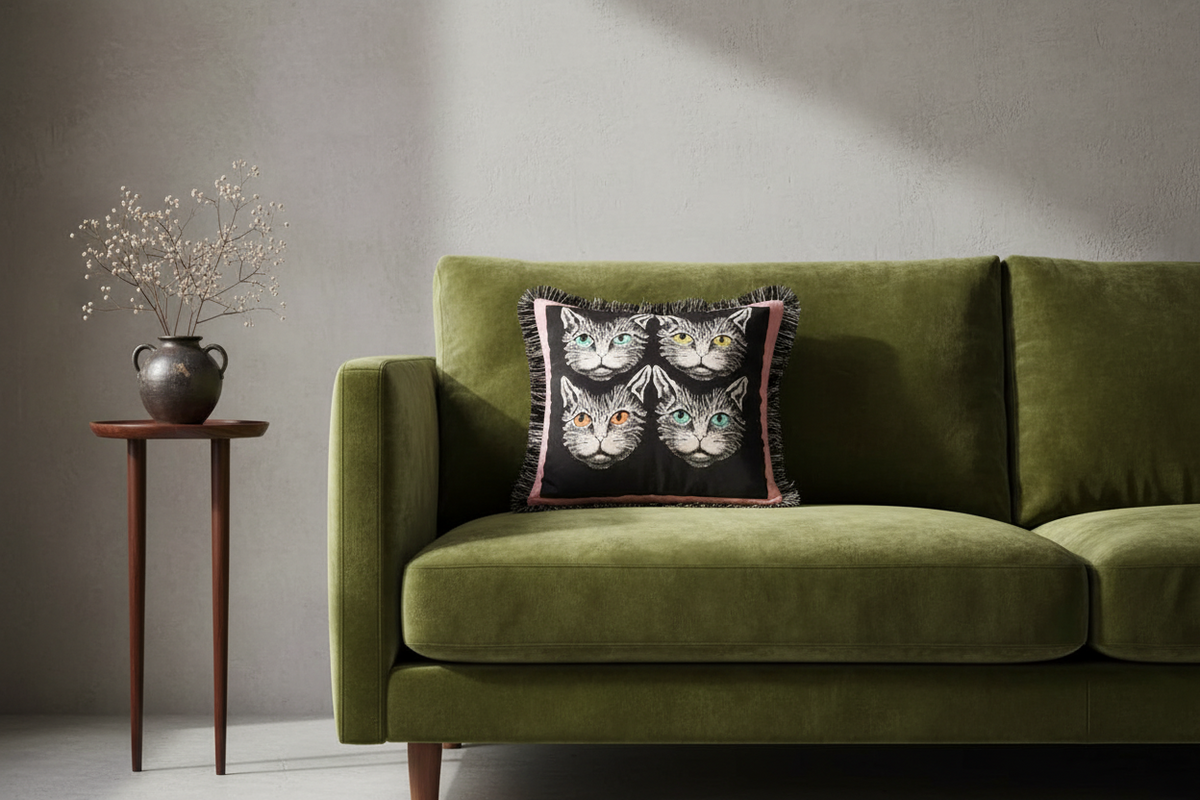 An olive green velvet sofa in a modern living room setting, styled with a square throw pillow featuring four illustrated cat faces on a black background with pink fringe.