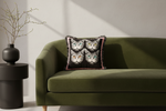 An olive green velvet sofa in a modern living room setting, styled with a square throw pillow featuring four illustrated cat faces on a black background with pink fringe.