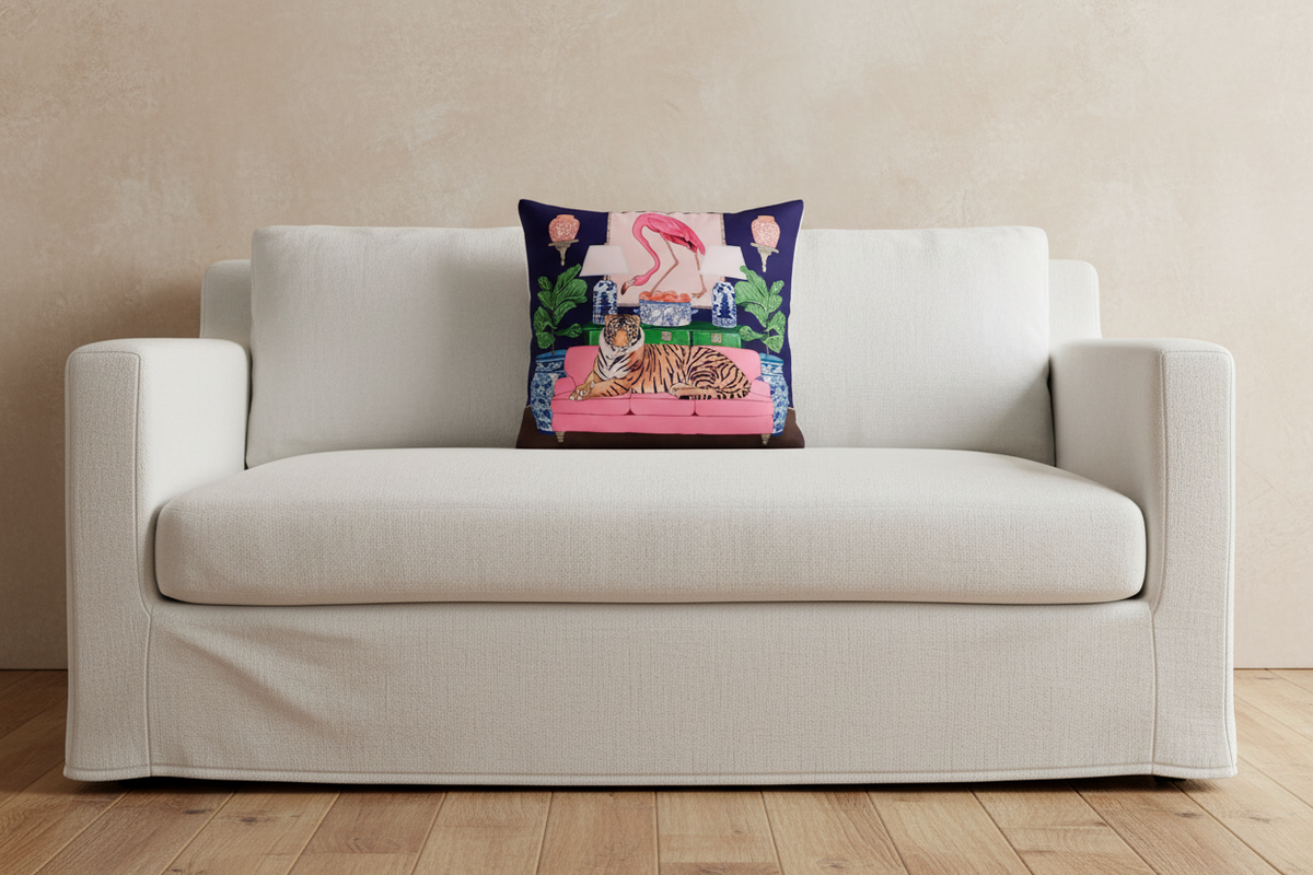 A square decorative throw pillow with a colorful tropical flamingo and tiger print, styled on a cream-colored fabric slipcovered sofa against a textured beige wall and light wood floor.