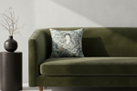 A square decorative throw pillow with a vintage-style woman illustration and floral pattern, resting on an olive green velvet sofa next to a black side table with a vase of branches.