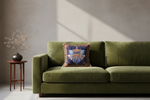 An olive green velvet sofa in a styled room setting, featuring a decorative blue and gold patterned throw pillow. A small wooden side table with a dark vase of dried branches stands to the left against a textured gray wall with soft diagonal sunlight shadows.