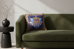 An olive green velvet sofa in a modern living room setting, featuring a square decorative throw pillow with a blue and gold heraldic shield design and fringed edges.