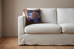 A cream-colored slipcovered sofa in a minimalist room with a light wood floor and beige wall, featuring a decorative blue and gold patterned throw pillow on the left cushion.