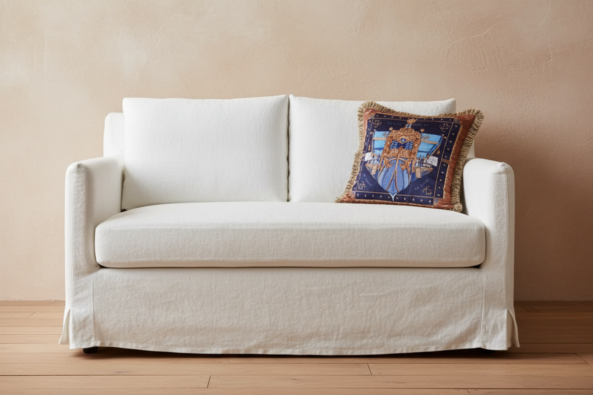 A white linen slipcovered loveseat sofa with a decorative blue and gold patterned accent pillow, set against a textured beige wall and light wood flooring.
