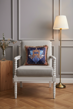 A decorative square throw pillow with a blue and gold heraldic crest design, featuring fringed edges, styled on a light gray upholstered armchair in a modern living room setting with a floor lamp and wood paneling.