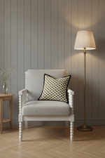 A light gray upholstered armchair with a black and gold patterned accent pillow, set in a room with a gray wood-paneled wall, a lit floor lamp, and a small wooden side table.