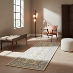 A long rectangular area rug with a beige and gray traditional floral pattern is styled in a modern, sunlit room with light wood floors, a pink armchair, a bench, a floor lamp, and a white pouf.