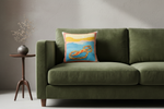 An olive green velvet sofa in a minimalist living room setting, styled with a square throw pillow featuring a colorful beach sunset illustration and a small wooden side table with a vase of dried branches.