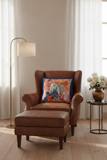 A brown leather wingback armchair with an attached ottoman in a bright, neutral room. A colorful floral throw pillow with a white horse design rests on the chair. A gold floor lamp stands to the left, and a small side table with white flowers is on the right.