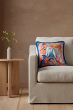 A square decorative throw pillow with a colorful horse and floral print, styled on a cream-colored armchair in a modern living room setting.