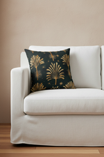A square throw pillow with a black background and gold palm tree pattern, styled on a white sofa against a beige wall.