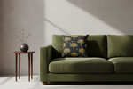 An olive green velvet sofa in a modern living room setting, styled with a dark green throw pillow featuring a gold palm tree pattern. A small wooden side table with a dark vase of dried branches stands to the left against a textured gray wall with diagonal sunlight shadows.