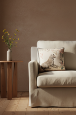 A white slipcovered sofa in a minimalist room with a textured beige wall and light wood flooring, featuring a decorative throw pillow with a leopard print.