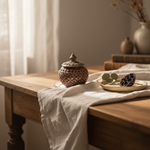 A small decorative ceramic lidded jar with an intricate geometric pattern sits on a draped white linen table runner on a rustic wooden table, styled with a small plate and eucalyptus leaves in a warm, softly lit room.