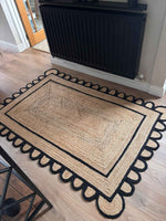 A beautifully crafted handmade black scalloped jute rug placed on a wooden floor in a cozy living room setting. This area rug, perfect for living room spaces, features a natural jute texture with elegant black scalloped edges, capturing the essence of rustic yet stylish design. Ideal for those looking for an 8x10 living room rug that adds warmth and character.