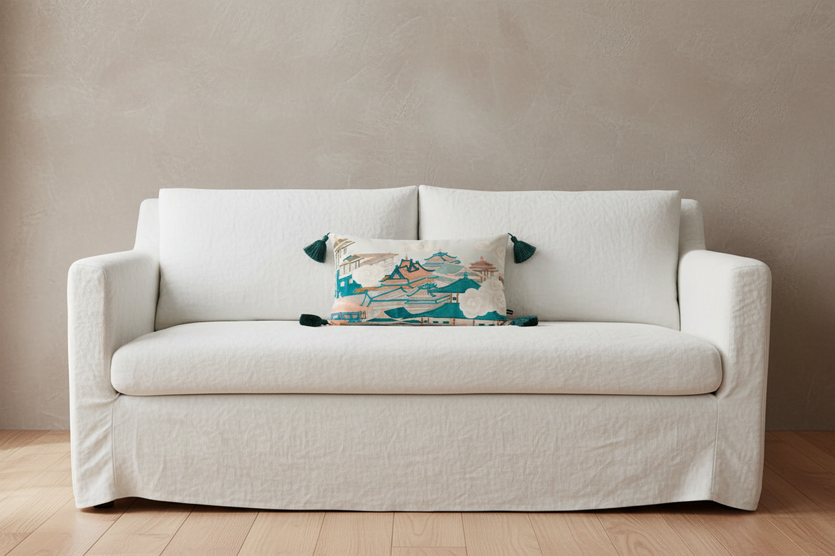 A cream-colored slipcovered loveseat sofa with a decorative patterned accent pillow, set against a textured beige wall and light wood flooring.