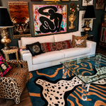 A styled living room featuring a white sofa with patterned throw pillows, a leopard-print armchair, a blue and tan abstract-patterned rug, and multiple framed artworks on a dark wall.